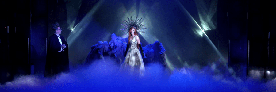 Usvan täyttämällä lavalla valojen keskellä kruunupäinen nainen, jota ympäröi kaapuihin puettu ihmisjoukko. Lavan sivulla frakkiin pukeutunut pystyhiuksinen ja silmälasipäinen mies