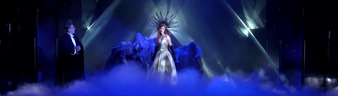 Usvan täyttämällä lavalla valojen keskellä kruunupäinen nainen, jota ympäröi kaapuihin puettu ihmisjoukko. Lavan sivulla frakkiin pukeutunut pystyhiuksinen ja silmälasipäinen mies