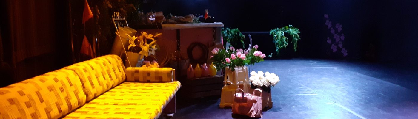 vasemmalla edessä vanha puurunkoinen, irtotyynyinen, kangassohva. Taustalla kukkakaupalavastus kukkineen tummasävyisessä ja hieman savuisessa teatteritilassa.