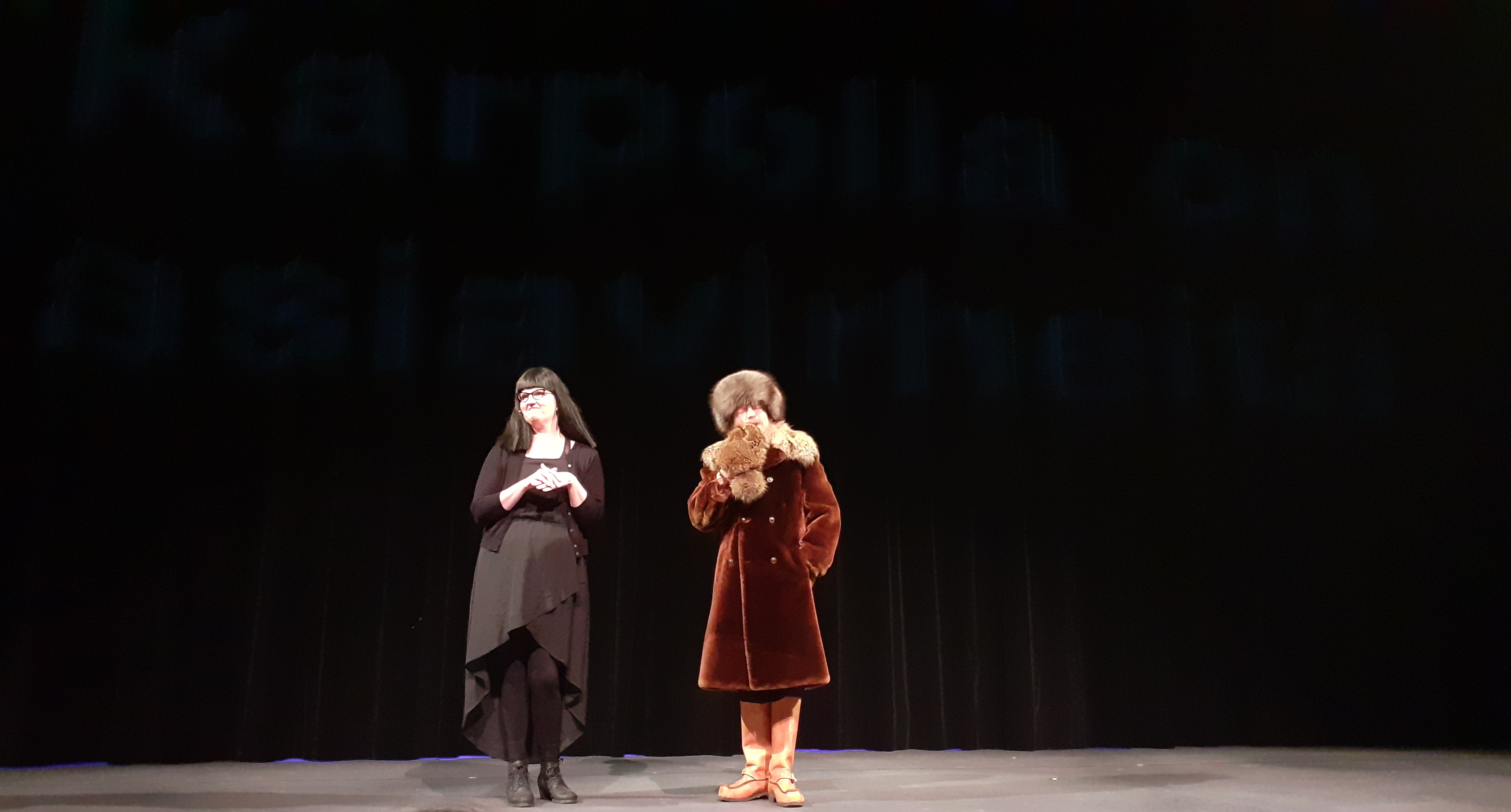 Teatterilavalla. Verhoihin heijastettu teksti "Karpolla on asiavirheitä" Vasemmalla nainen, jolla on pitkät mustat hiukset ja musta asu. Oikealla karvalakkiin ja takkiin pukeutunut Hannu Karpo -hahmo