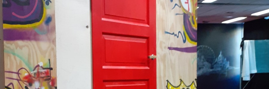 keskellä on punainen ovi, jonka molemmin puolin on graffitein koristellut levyseinät. Oikealla avautuu näkymä tilaan, jonka takaosassa on liitutaululle piirretty Helsingin rantaviiva.