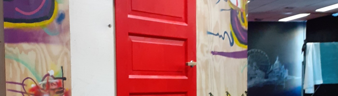 keskellä on punainen ovi, jonka molemmin puolin on graffitein koristellut levyseinät. Oikealla avautuu näkymä tilaan, jonka takaosassa on liitutaululle piirretty Helsingin rantaviiva.
