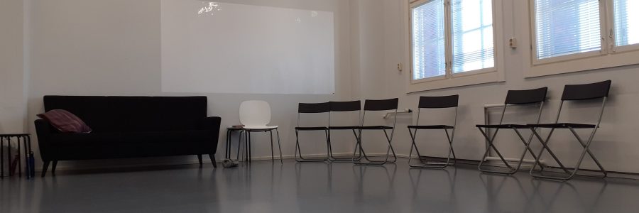 Nurkkaus tilasta. Vasemmalla musta sohva. Oikealla seinustalla, ikkunoiden alla, useita tuoleja rivissä seinän vierustalla.