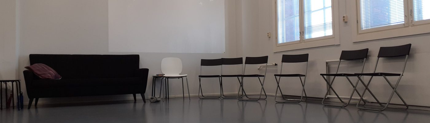 Nurkkaus tilasta. Vasemmalla musta sohva. Oikealla seinustalla, ikkunoiden alla, useita tuoleja rivissä seinän vierustalla.