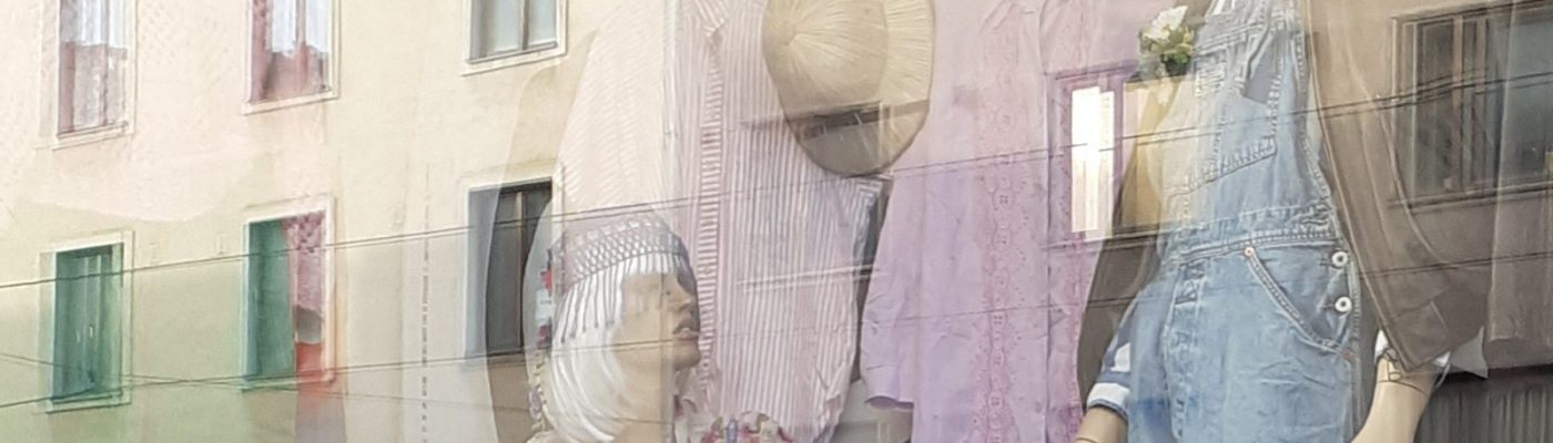 Näyteikkuna, jossa nais- ja miesmannekiini nukke vanhanaikaisiin asuihin puettuna. Ikkunasta heijastuu auringonpaiste vastapäisen kerrostalon ikkunoissa.