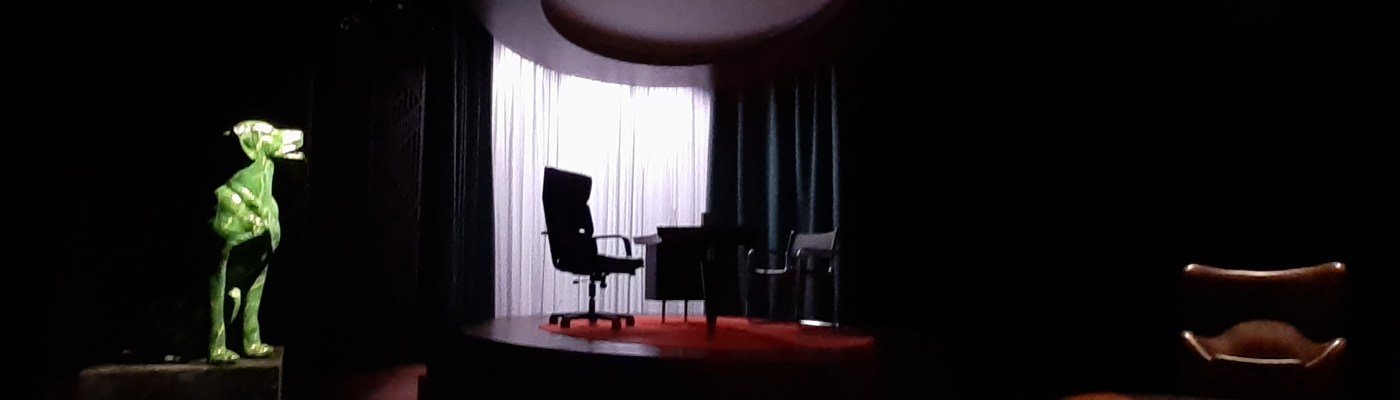 Hämärä teatterin näyttämö. Vasemmalla valokeilassa vihreä koirapatsas. Keskellä korokkeella konttoripöytä ja kaksi tuolia. Oikeassa laidassa ruskea nahkatuoli.