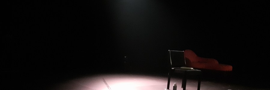 Musta lava, jonka keskellä valo valaisee mustan tuolin, jonka alla on juomapullo. Taustalla näkyy punainen divaani.