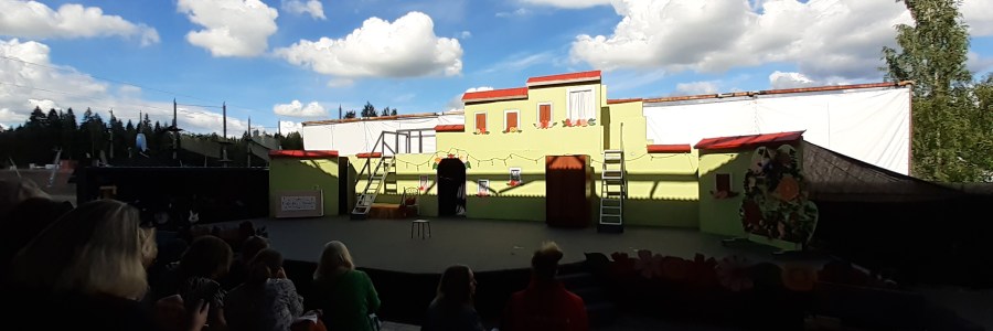 Kesäteatterin lava, jossa on lavasteena kaksikerroksinen rakennus. Taustalla näkyy poutapilvinen taivas.