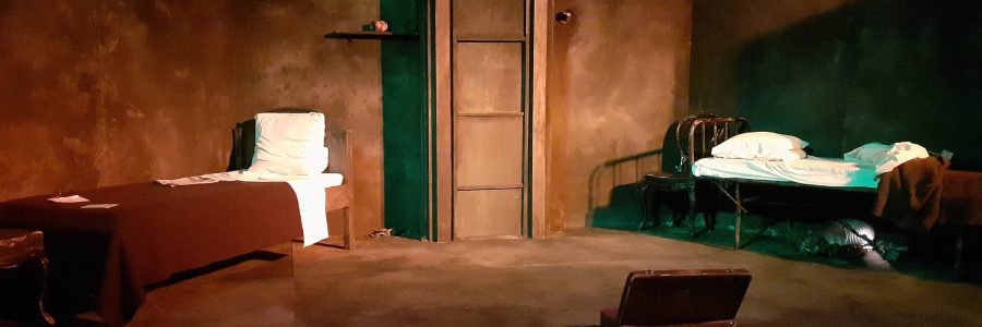 Etualalla avattu vanha matkalaukku. Taustalla kaksi diagonaaliin asetettua sänkyä, joiden välissä on ruokahissin muodostama ulkonema. Vasemman puoleinen sänky on siististi pedattu. Oikeanpuoleinen sänky, jonka takana näkyy myös tuoli, on avattu.