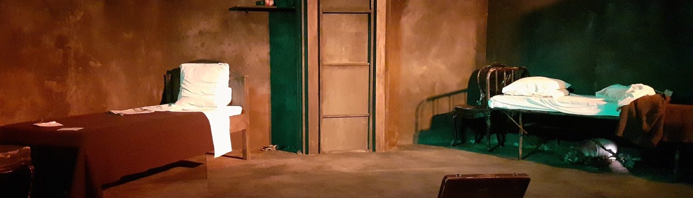 Etualalla avattu vanha matkalaukku. Taustalla kaksi diagonaaliin asetettua sänkyä, joiden välissä on ruokahissin muodostama ulkonema. Vasemman puoleinen sänky on siististi pedattu. Oikeanpuoleinen sänky, jonka takana näkyy myös tuoli, on avattu.