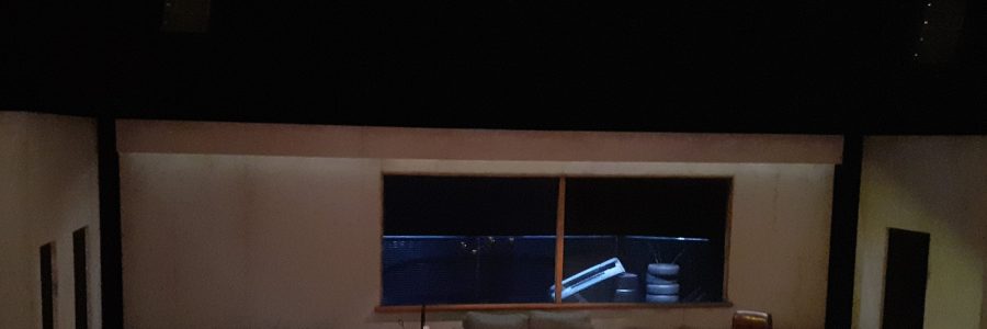 Laaja näyttämötila, jossa on taustalla valtavat ikkunat. Keskellä tilaa sohvapöytä, sohva, matto ja pyörivä nojatuoli.