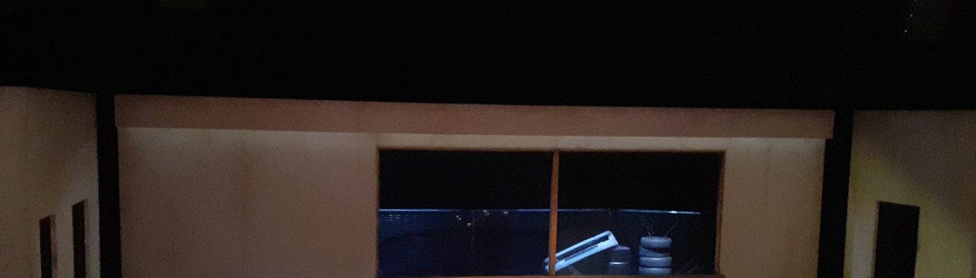 Laaja näyttämötila, jossa on taustalla valtavat ikkunat. Keskellä tilaa sohvapöytä, sohva, matto ja pyörivä nojatuoli.