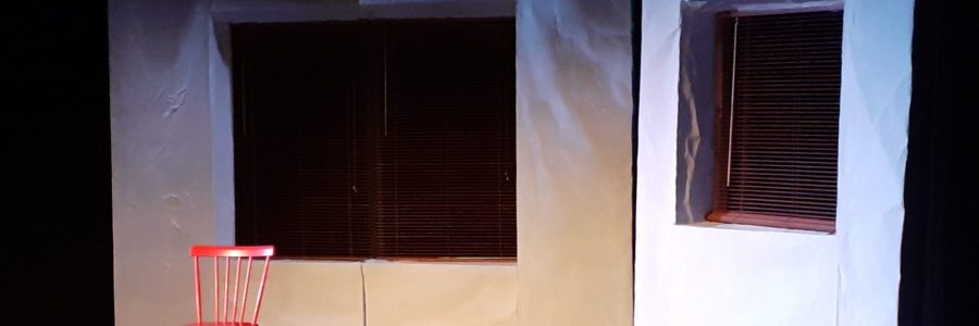 Valkoiset lavasteseinät, joiden keskellä on sälekaihtimin varustetu ikkunat. Etualalla vanha radio ja kirkkaan värinen puutuoli.