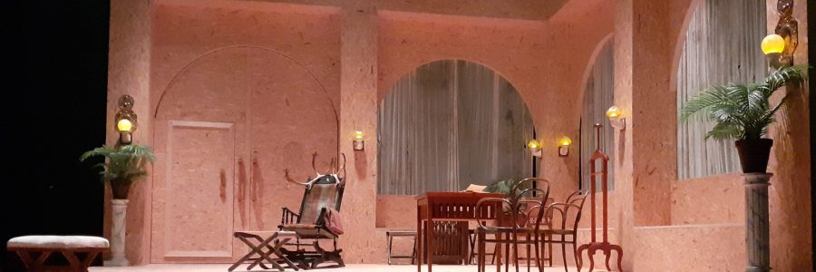 Lämpimän värinen teatterilavastus vanhahtavasta tilasta, jossa on puisia kalusteita, huonekasveja pilarien päällä sekä seinissä lampetteja.