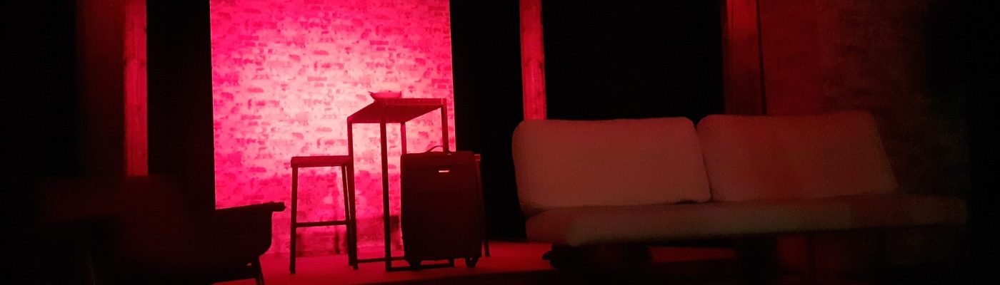 Punasävyinen lavastus, jossa etualalla on sohvatuoli ja nojaton sohva. Keskellä takana korkea pöytä, baarituoli ja matkalaukku.