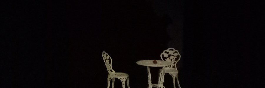 Tumman taustan keskellä valkoinen, metallinen, puutarhapöytä ja kaksi puutarhatuolia.