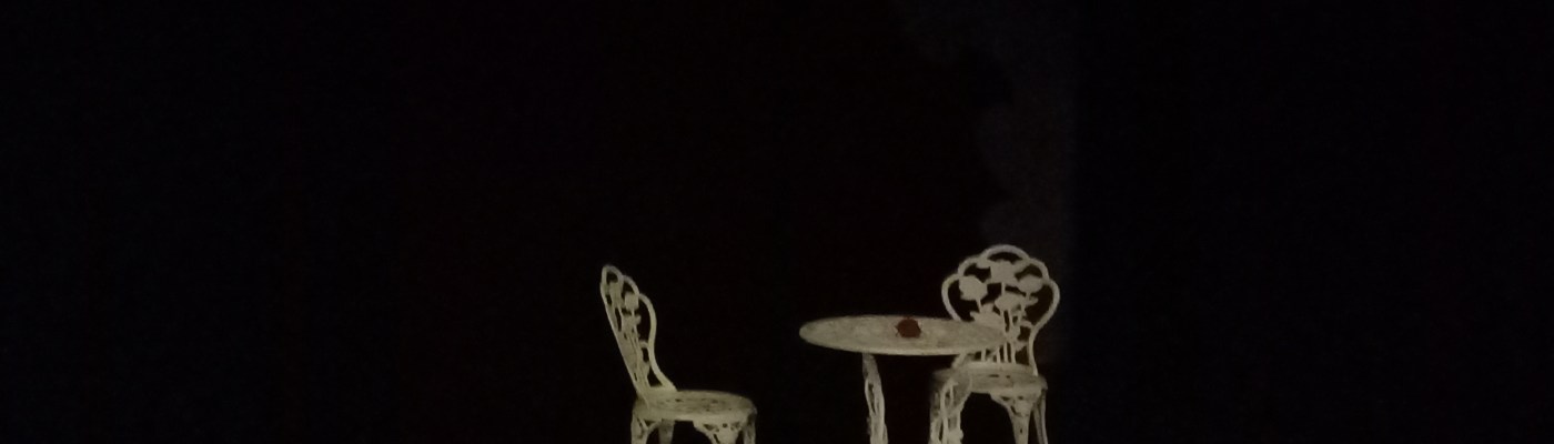 Tumman taustan keskellä valkoinen, metallinen, puutarhapöytä ja kaksi puutarhatuolia.