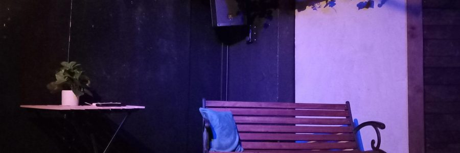 Ulkonäkymä teatterilavalla. Ulkotila, jossa pieni pöytä ja penkki. Penkin yllä on villiviinikehikko.