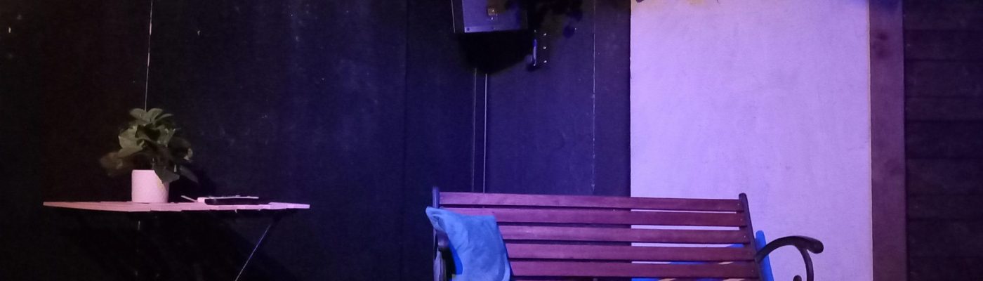 Ulkonäkymä teatterilavalla. Ulkotila, jossa pieni pöytä ja penkki. Penkin yllä on villiviinikehikko.