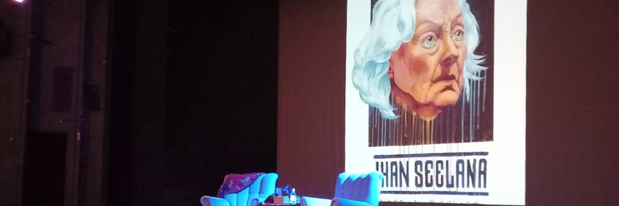 Punertavan sävyinen lavastus teatterissa. Lavan keskellä kaksi nojatuolia sekä pieni pöytä. Etualalla punainen matto. Taustalla heijastuksena Seela Sellan piirroskuva sekä teksti "Ihan Seelana".