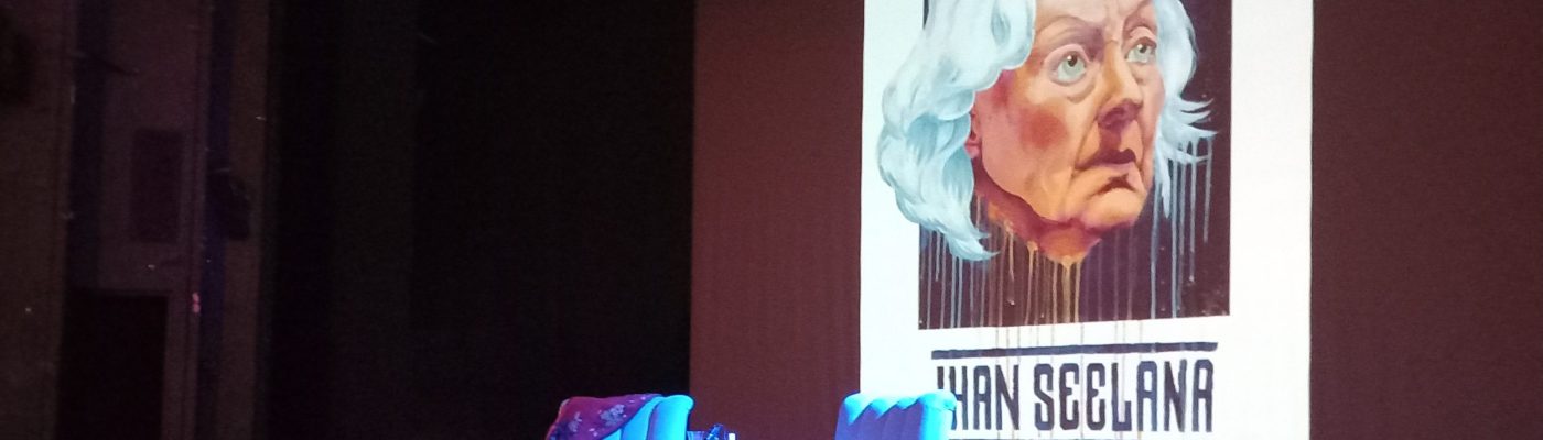 Punertavan sävyinen lavastus teatterissa. Lavan keskellä kaksi nojatuolia sekä pieni pöytä. Etualalla punainen matto. Taustalla heijastuksena Seela Sellan piirroskuva sekä teksti "Ihan Seelana".