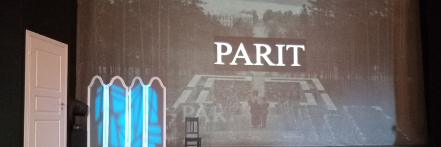 Kuvassa teatterilavassa, jossa vasemmassa laidassa on valkoinen ovi. Taustalle on projisoitu mustavalkokuva Jyväskylästä, Neron portailta Jyväsjärvelle päin. Taustakuvan päällä ok teksti "parit". Lisäksi lavalla on pukeutumissermi ja yksi tuoli.