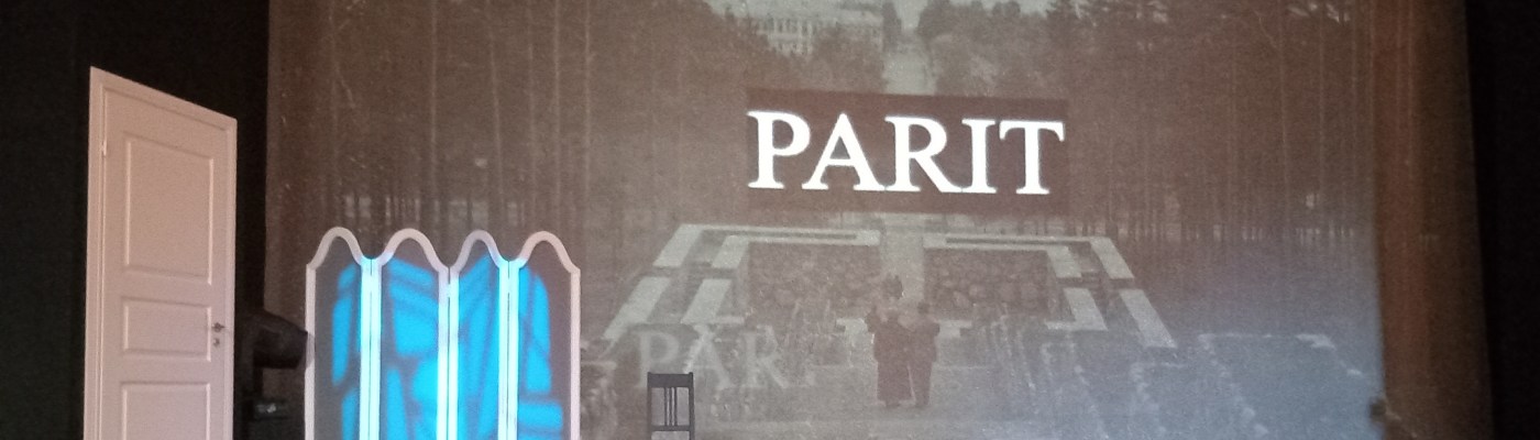 Kuvassa teatterilavassa, jossa vasemmassa laidassa on valkoinen ovi. Taustalle on projisoitu mustavalkokuva Jyväskylästä, Neron portailta Jyväsjärvelle päin. Taustakuvan päällä ok teksti "parit". Lisäksi lavalla on pukeutumissermi ja yksi tuoli.