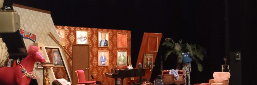 Teatterinäyttämö. Lavan reunalla pinkki hevonen. Keskialalla vanhahtava olohuonekalusto ja flyygeli.