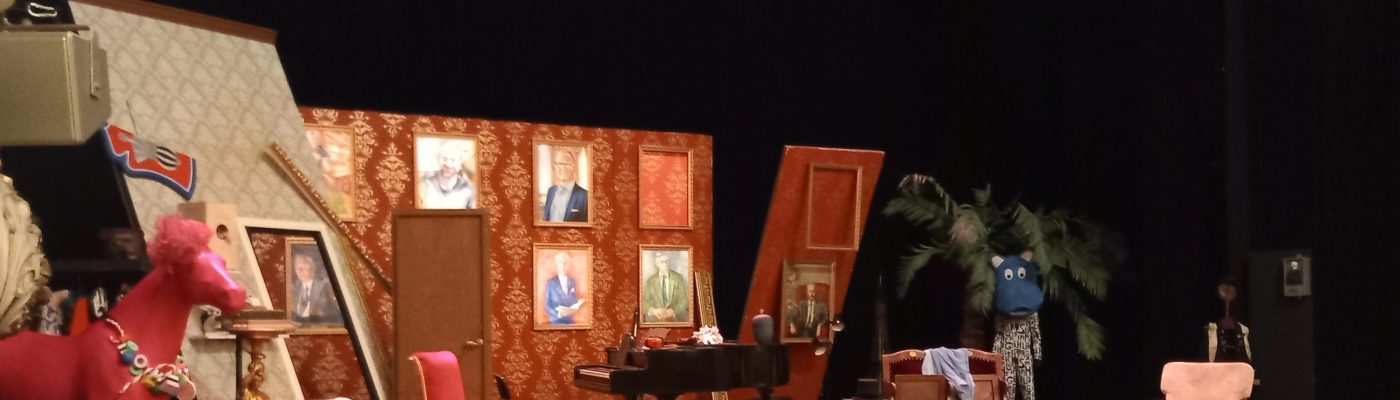 Teatterinäyttämö. Lavan reunalla pinkki hevonen. Keskialalla vanhahtava olohuonekalusto ja flyygeli.