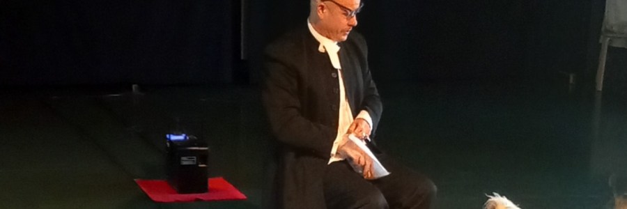 Näyttämöllä tummaan pukuun pukeutunut näyttelijä istumassa tuolilla ja katselemassa vieressään lattialla istuvaa pientä valkoista koiraa.