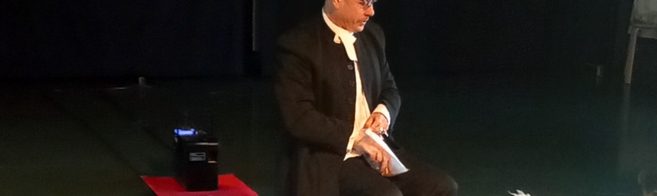 Näyttämöllä tummaan pukuun pukeutunut näyttelijä istumassa tuolilla ja katselemassa vieressään lattialla istuvaa pientä valkoista koiraa.